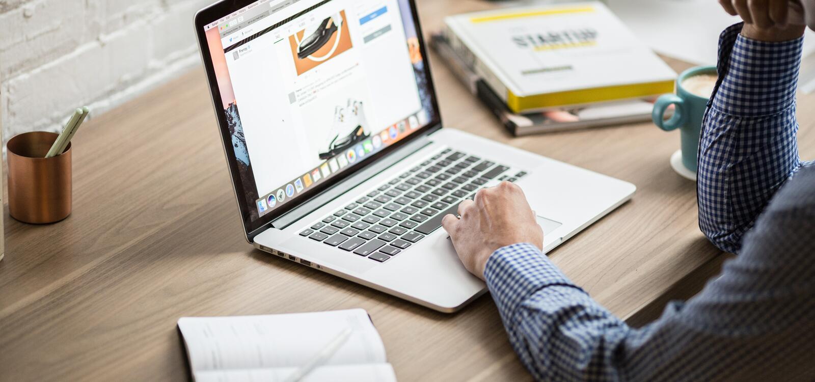 A close up of a man in a blue checkered shirt on the computer sittting at a desk and writing things down