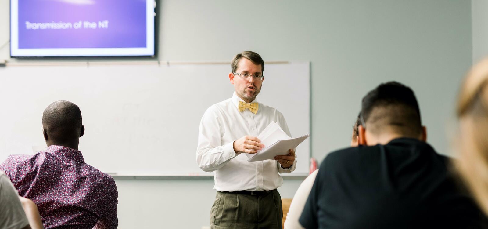 Dr. Campbell, dressed in a white buttoned shirt and yellow bow-tie, stands in front of his class and holds papers as he begins to hand them out to his students