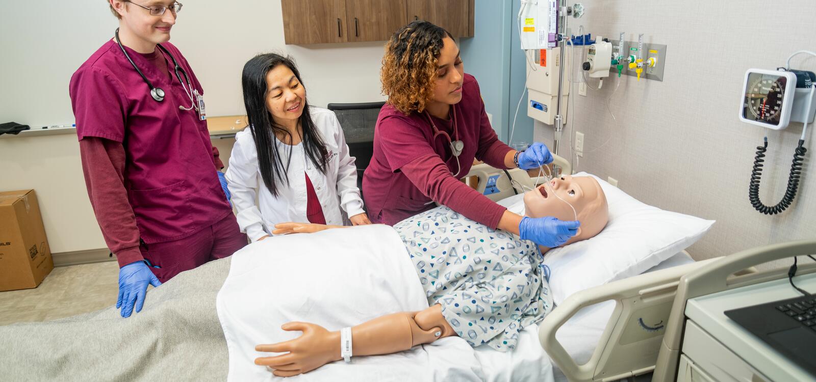 A nursing student, dressed in maroon scrubs, places oxygen tubes in a fake patients nose while a peer and professor stand by and watch