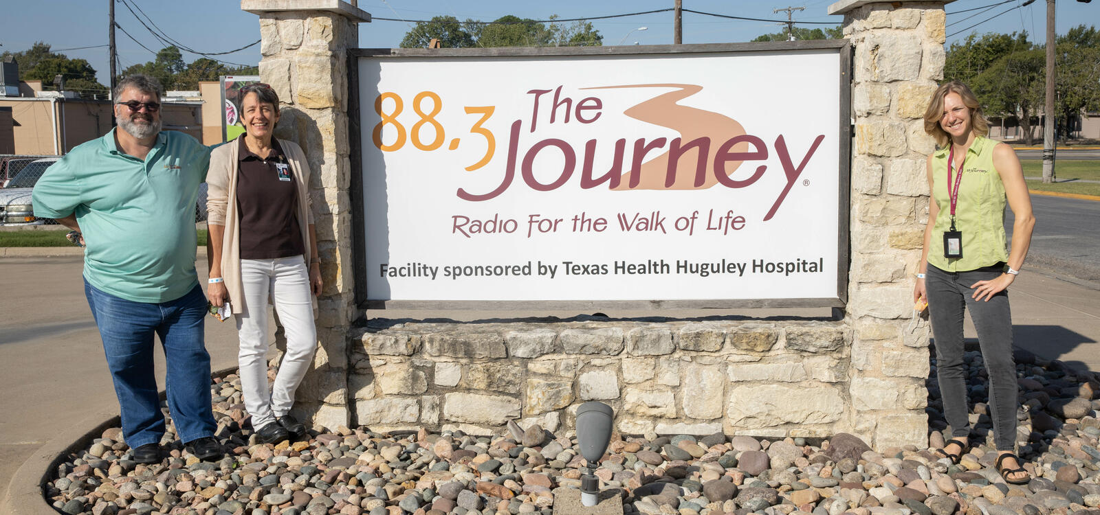 Mike and Wanda Agee stand to the left of the radio station sign that reads "88.3 The Journey, Radio for the Walk of Life." Danae stands to the right of the sign.