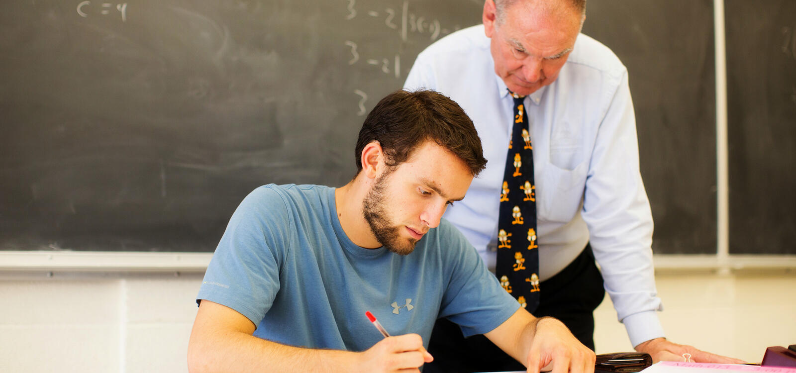 A student sits at a desk and completes his homework as his professor stands behind him to help