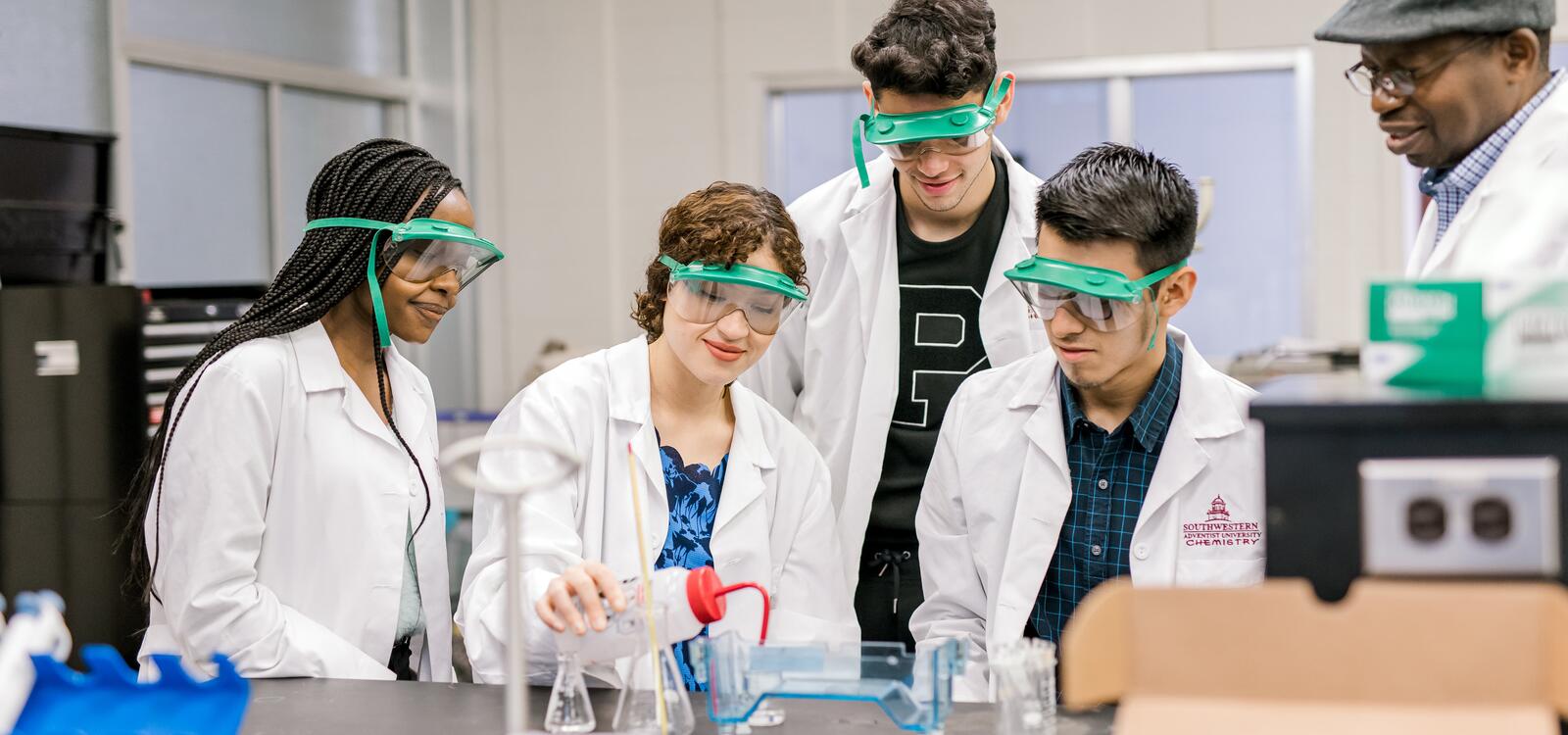 with help from their professor, three students, dressed in white lab coats and goggles, sit around and another student conduct an experiment. 