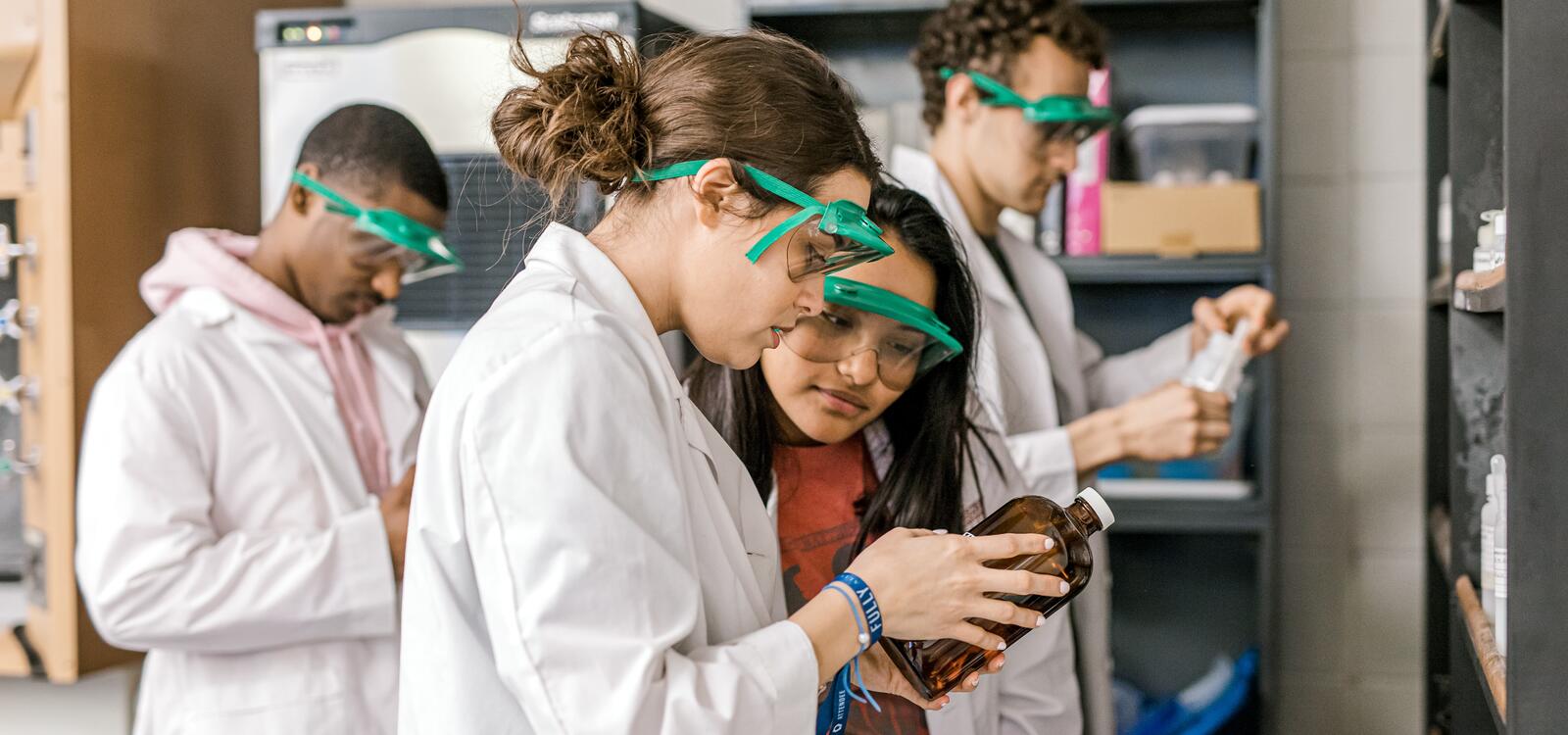 Two students, dressed in lab coats and goggles, pick up a bottle to read the label of the chemical