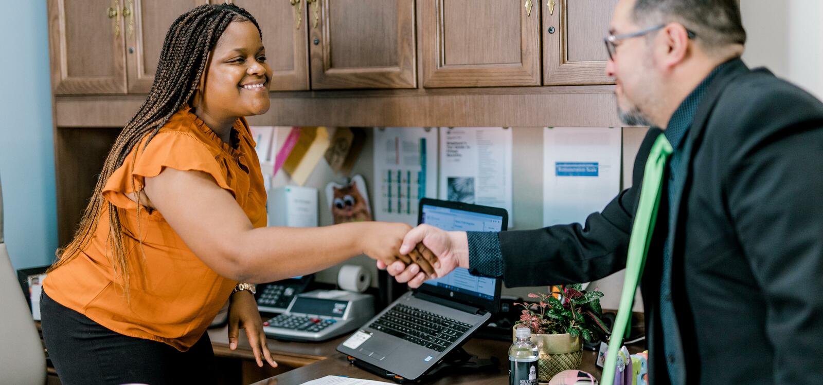 A female stands up from behind her desk and smiles as she leans forward to shake the hand of a gentleman standing on the other side of her desk