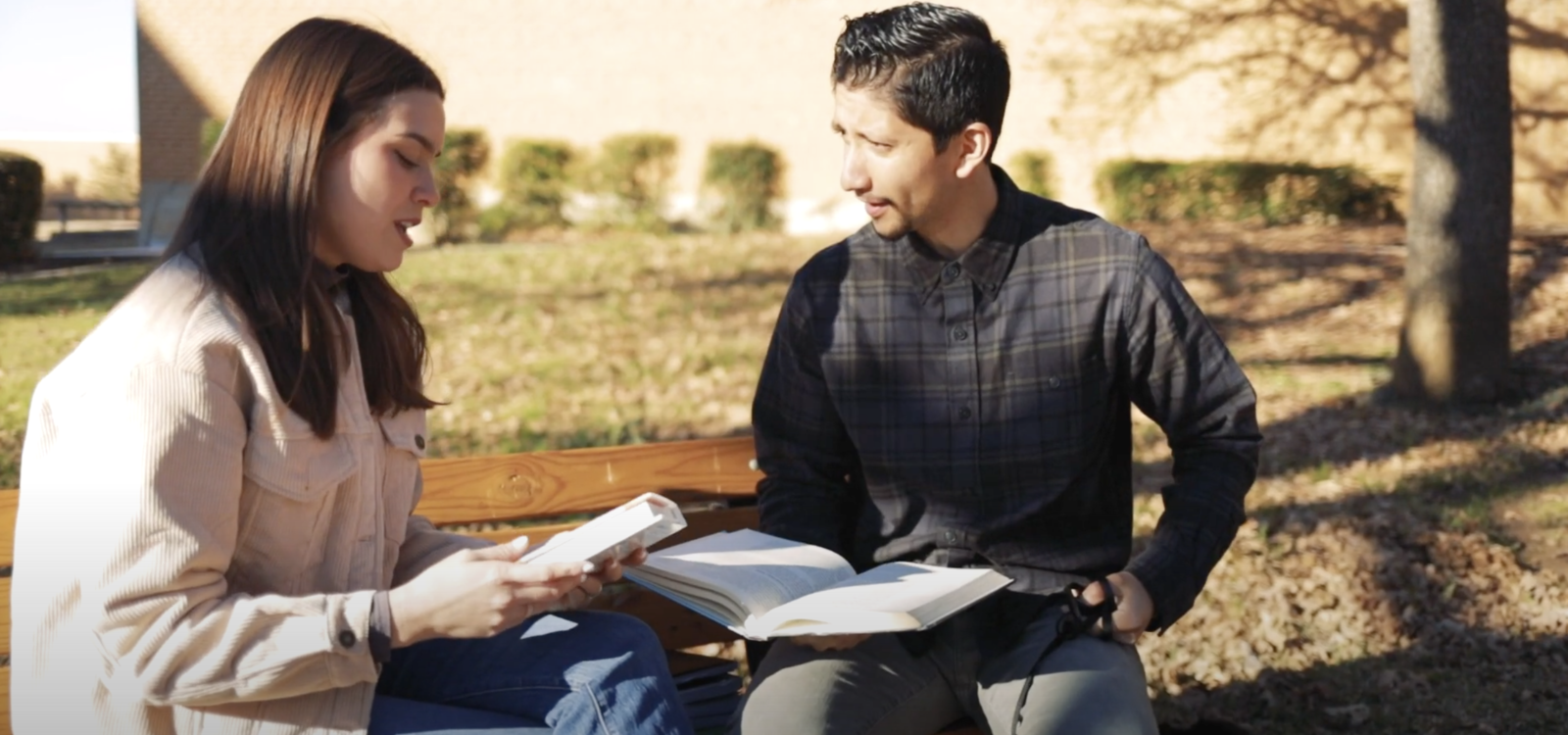 Two students sit on a bench holding books and talking