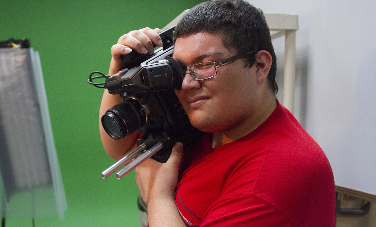 A young man squints one eye as he uses the other to look through the lense of a video camera