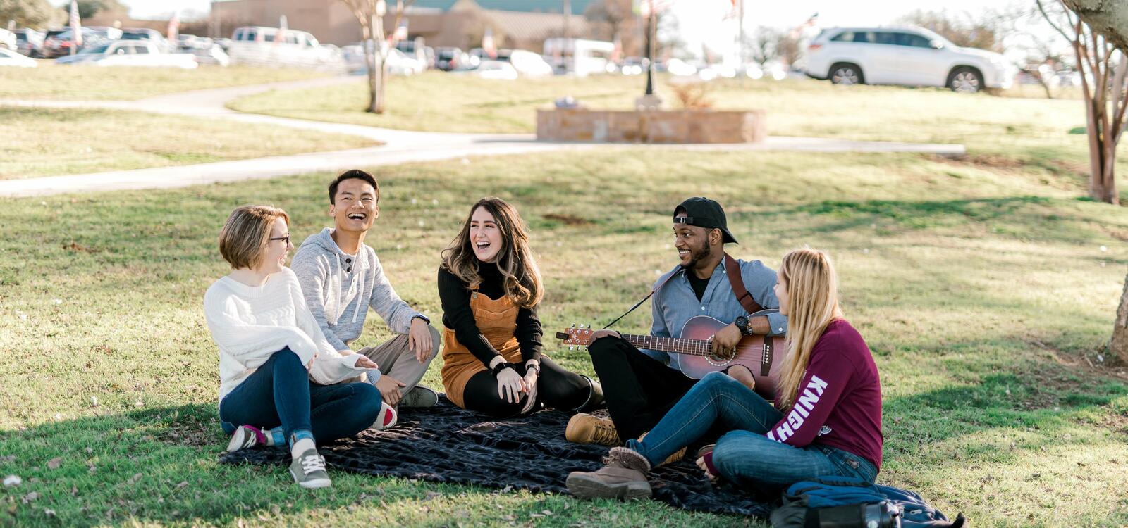 Five students sit under a tree on a blanket laughing and singing as one studentplays the guitar