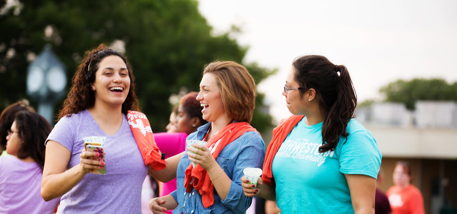 Three female students with snow cones in hand, smile and laugh as they stand outside