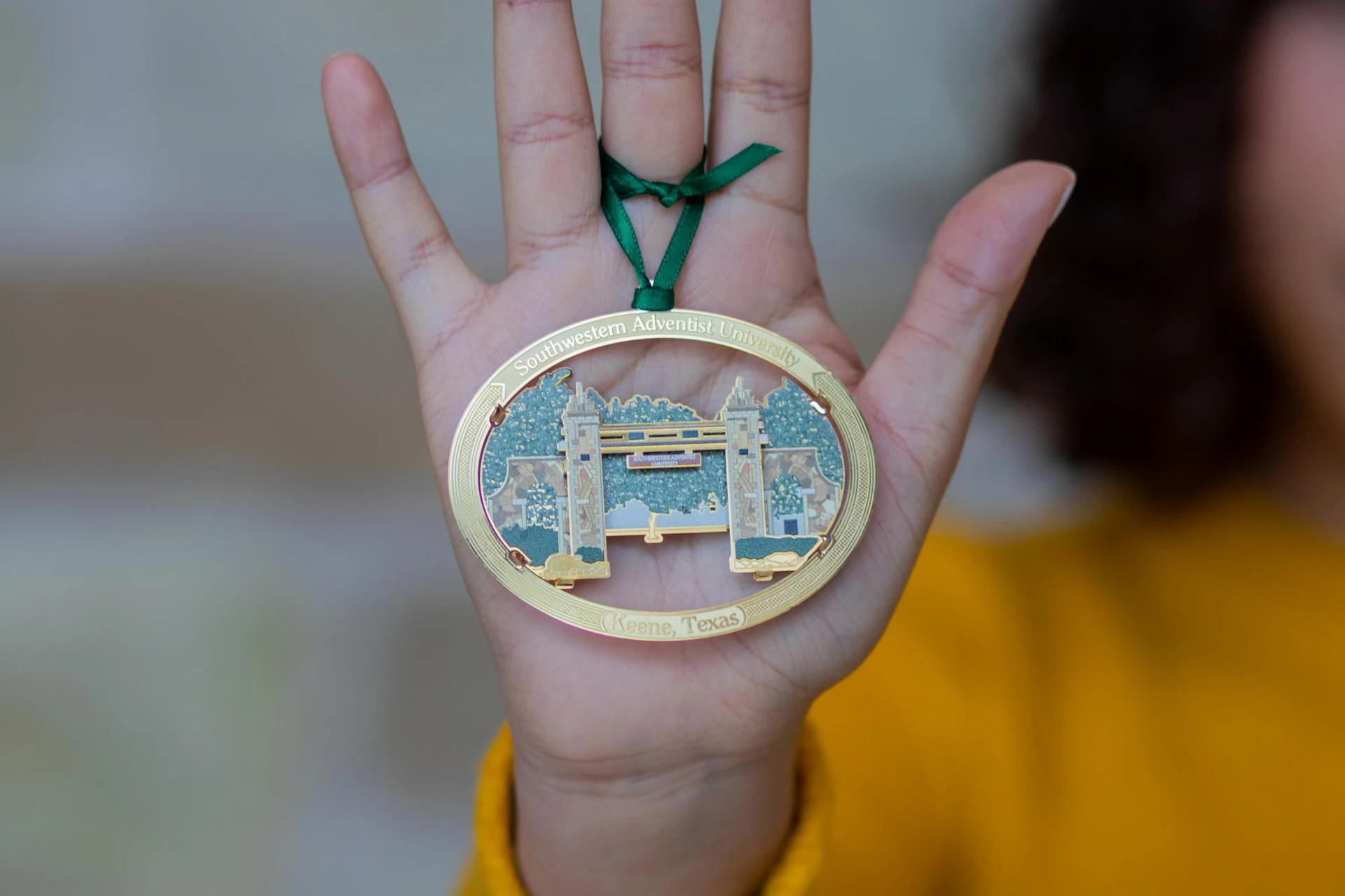 Oval ornament with "Southwestern Adventist University" at the top and "Keene, TX" at the bottom with the Mizpah gate and its surrounding trees in the center