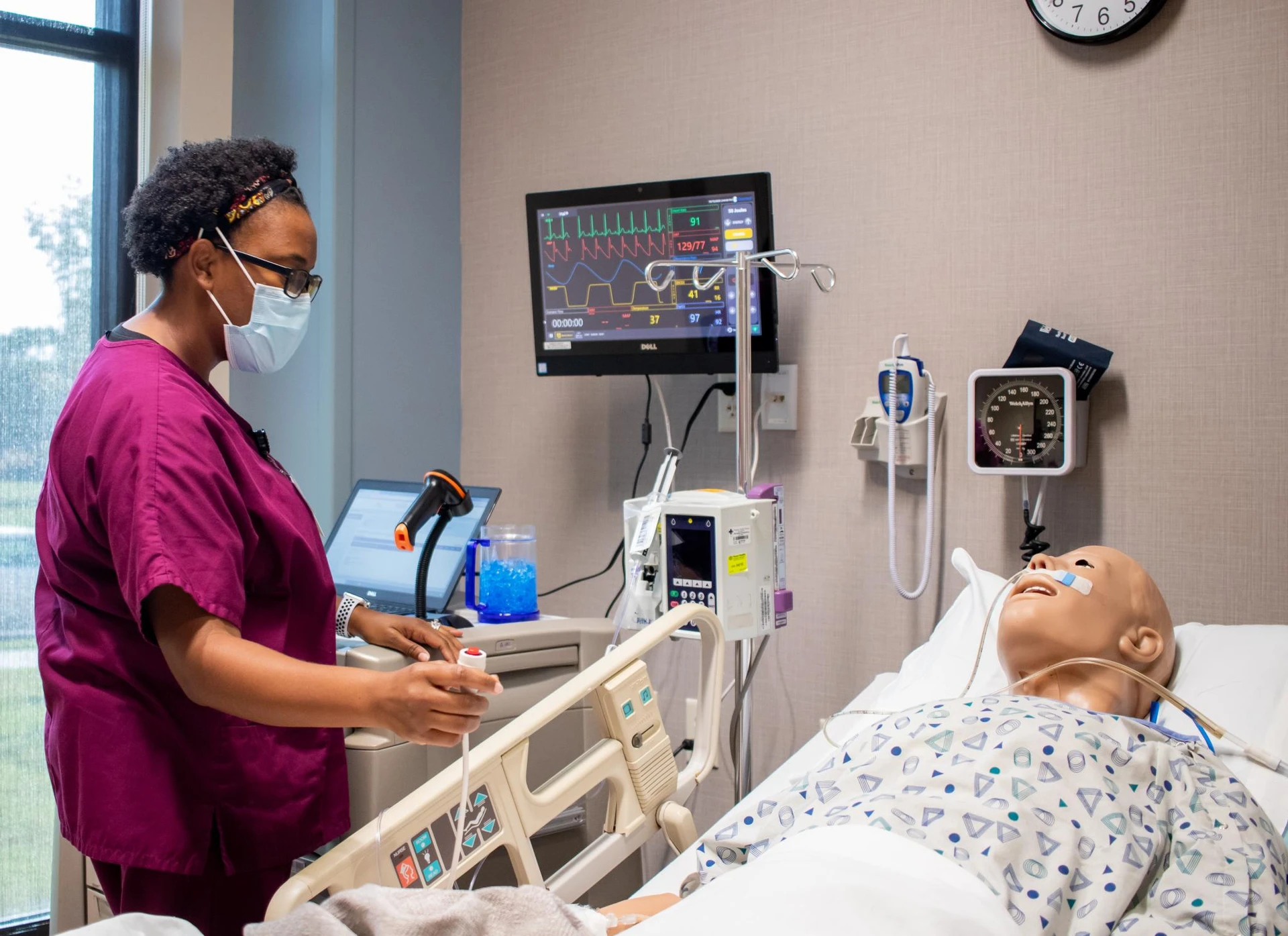 Female student in maroon scrubs checking vitals on a simulation mannequin.  