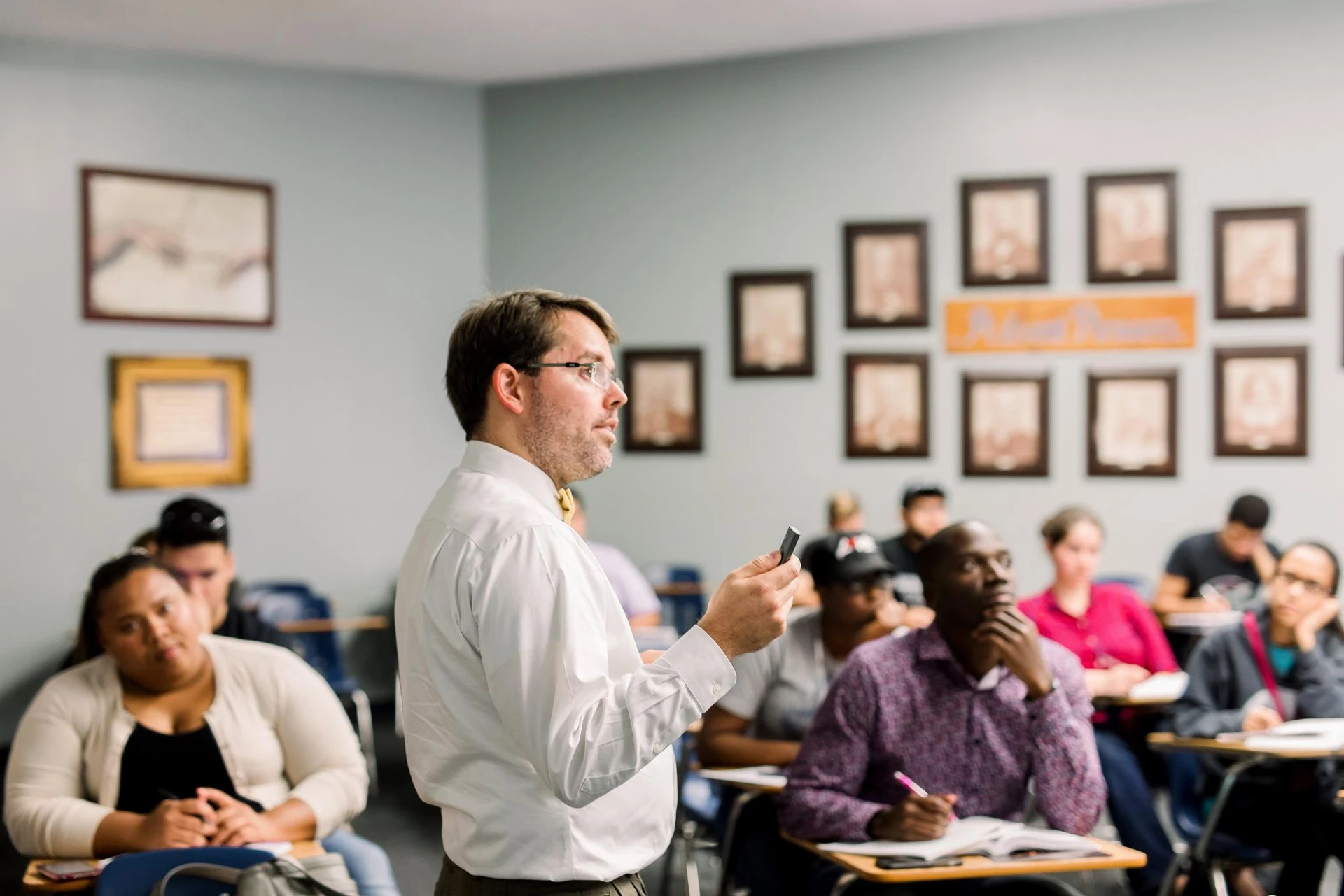 A professor, dressed in a white button up and yellow bowtie, looks towards a screen with a clicker in his hand