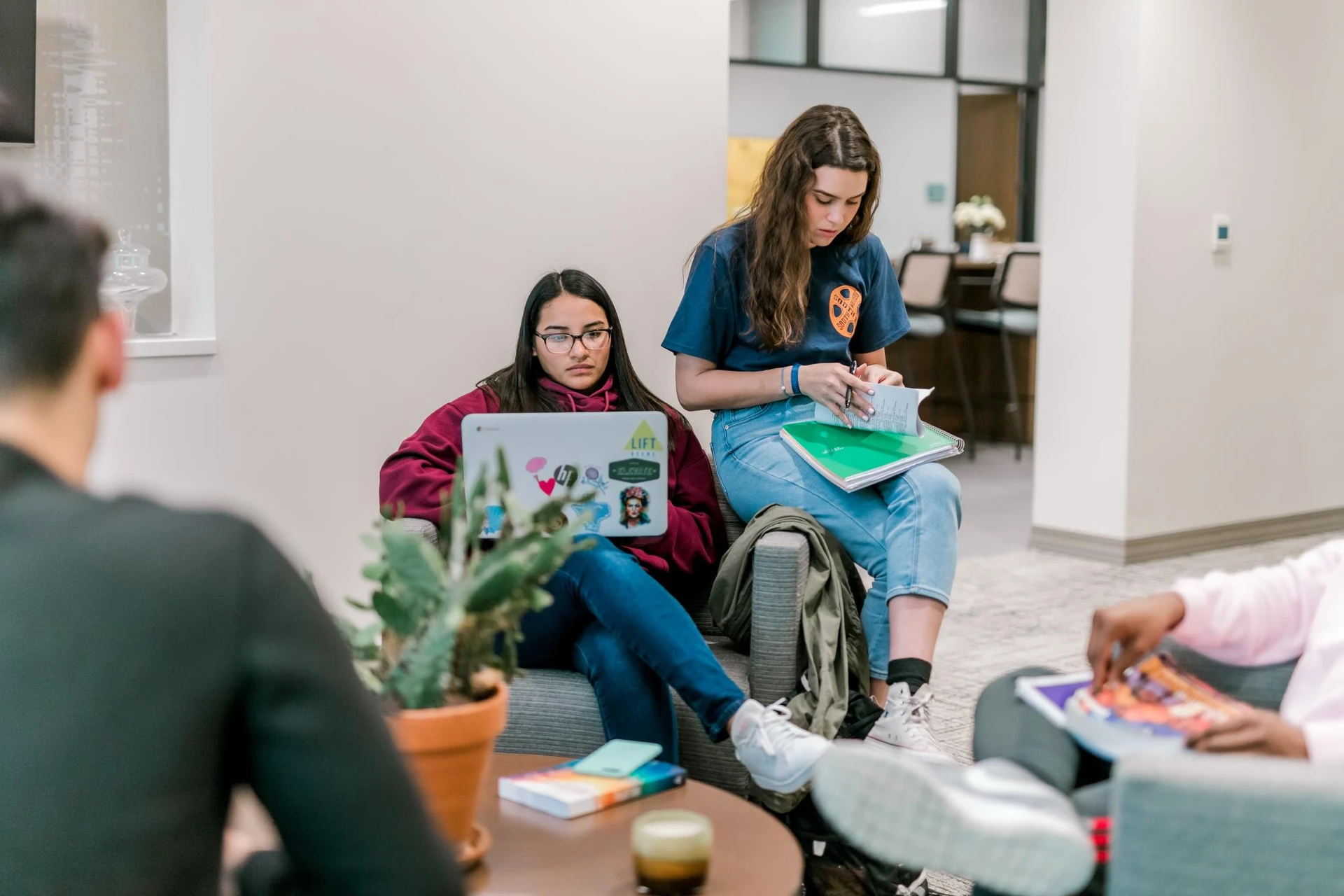 A female student sits in a chair as she complete asignments on her laptop, and another student sits on the arm of the chair as she opens up her notes