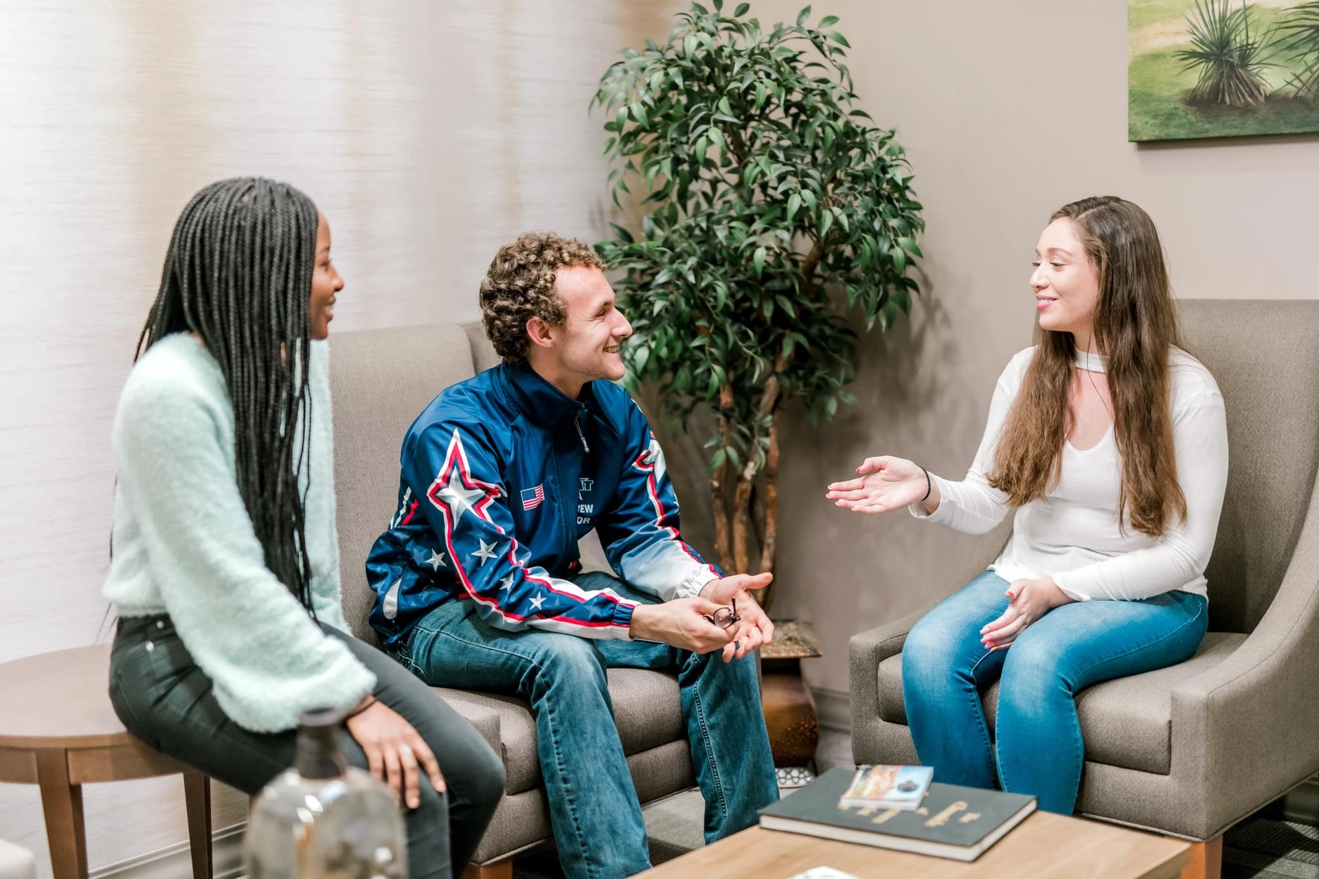 Two students sit in gray chairs, and another sits on a side table, as they talk