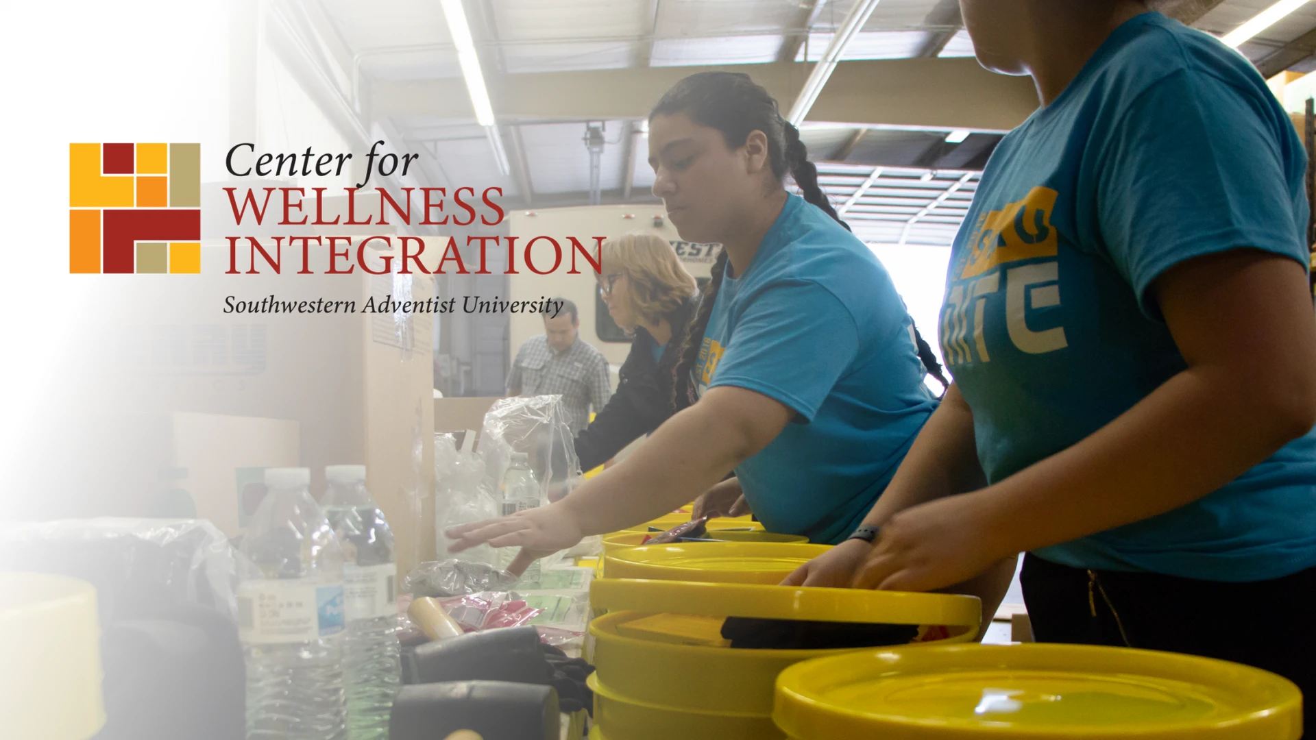 A group of students stand in a line and fill the yellow buckets in front of them with different necessary items, such as water, blankets and more.