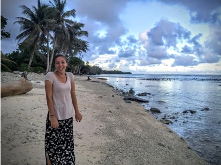 A woman, dressed in a white blouse and long skirt, smiles as she stand in front of the beautiful scenary at the beach