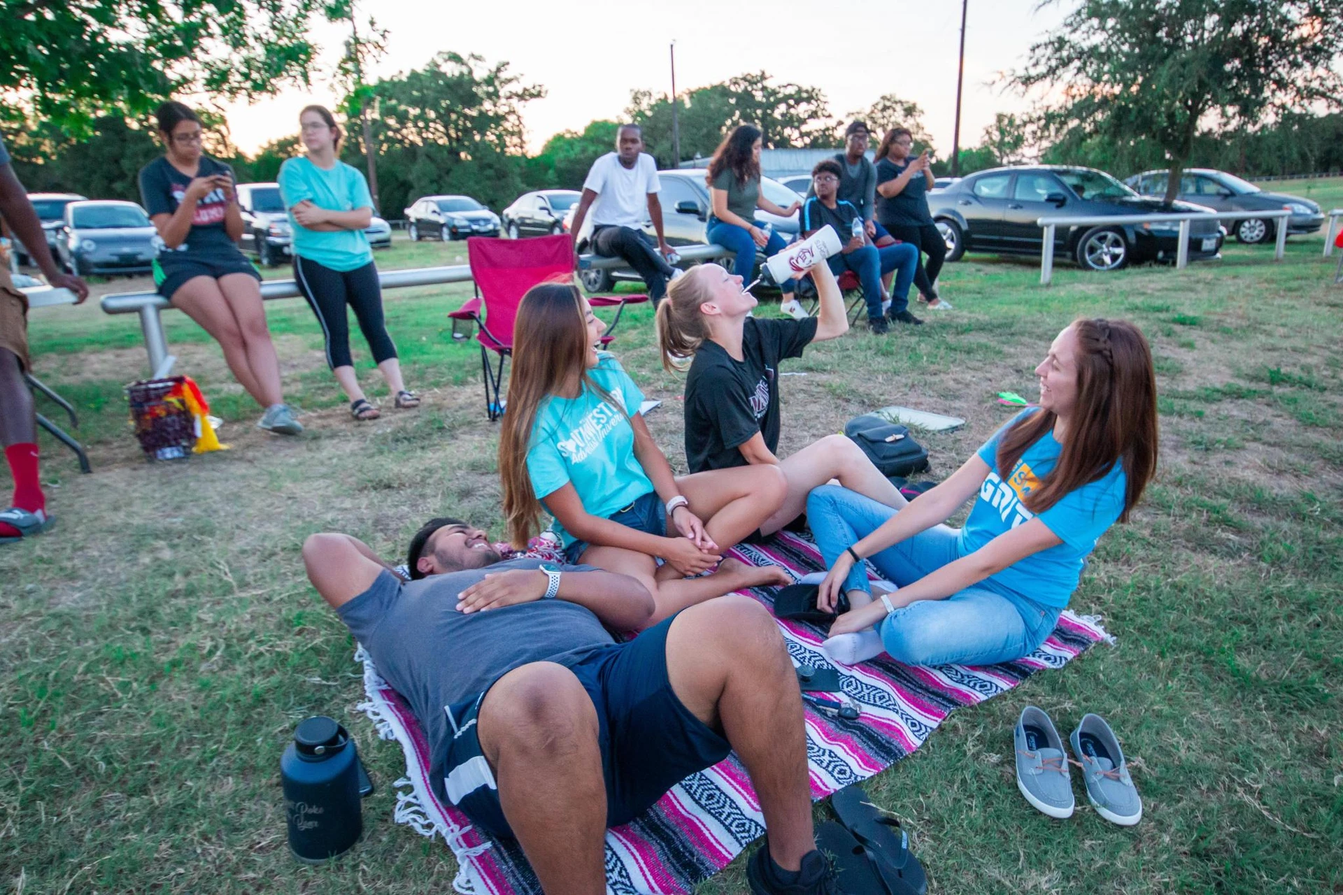 Watching outdoor activities, a group of four friends laugh and smile as they sit on an outdoor blanket