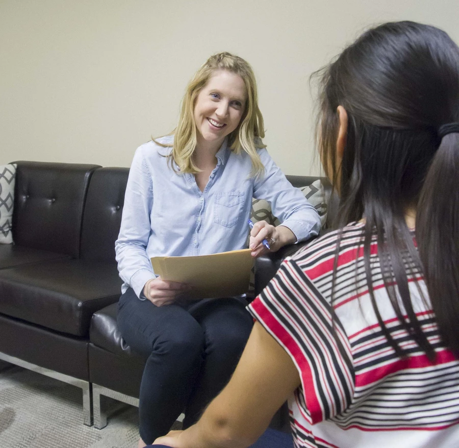 A blonde female smiles, holding a manilla folder and a pen, as she sits in front a brunette female