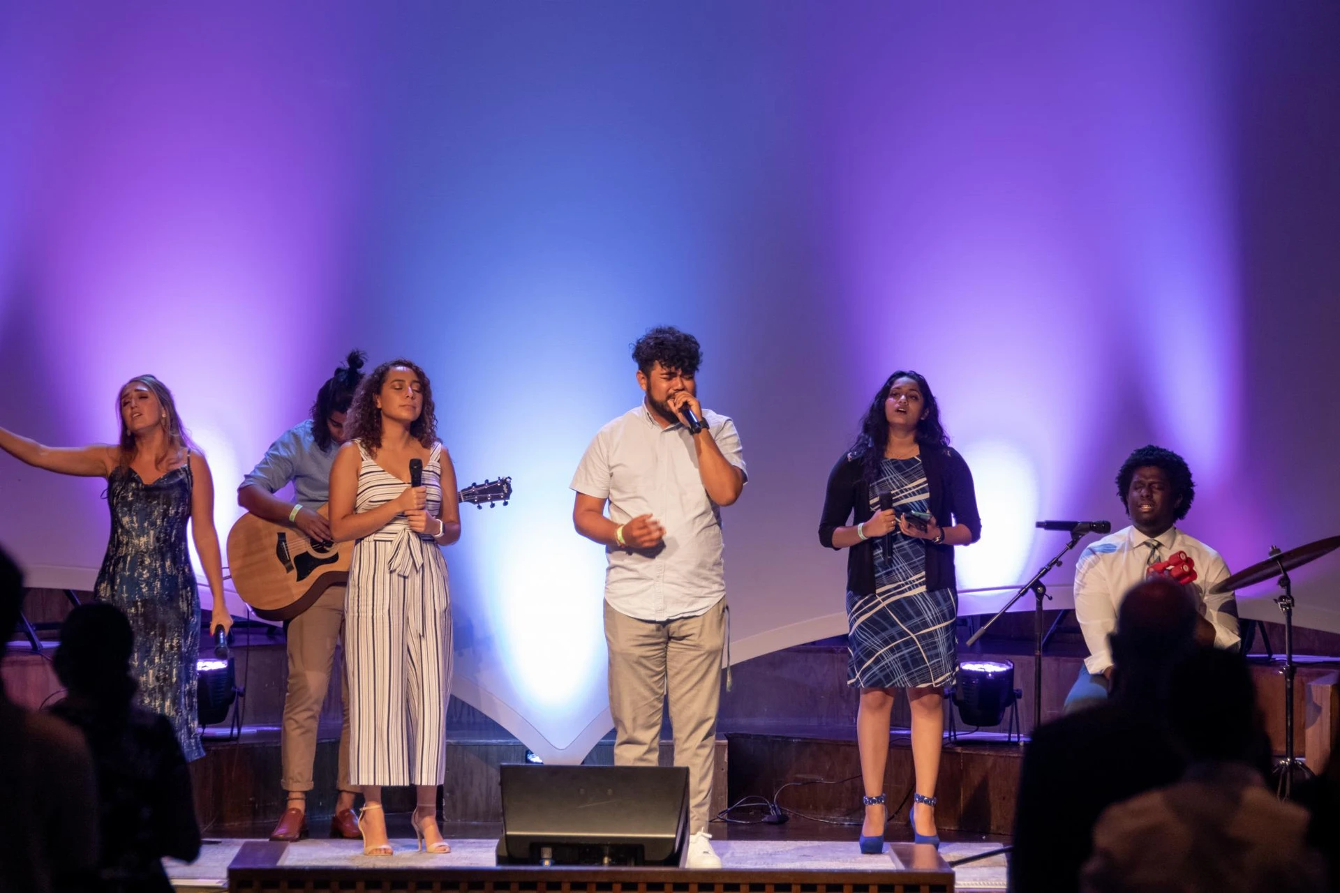 Four students stand on stage singing while one student stands in the back playing guitar and another sits on the right playing the cajon