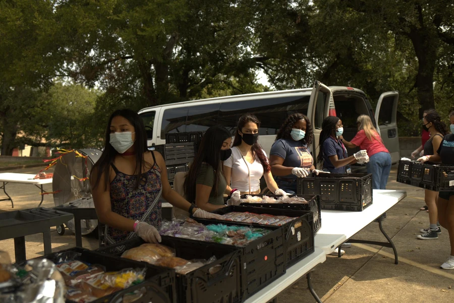 Students wearing masks and gloves stand behind tables with containers of food 