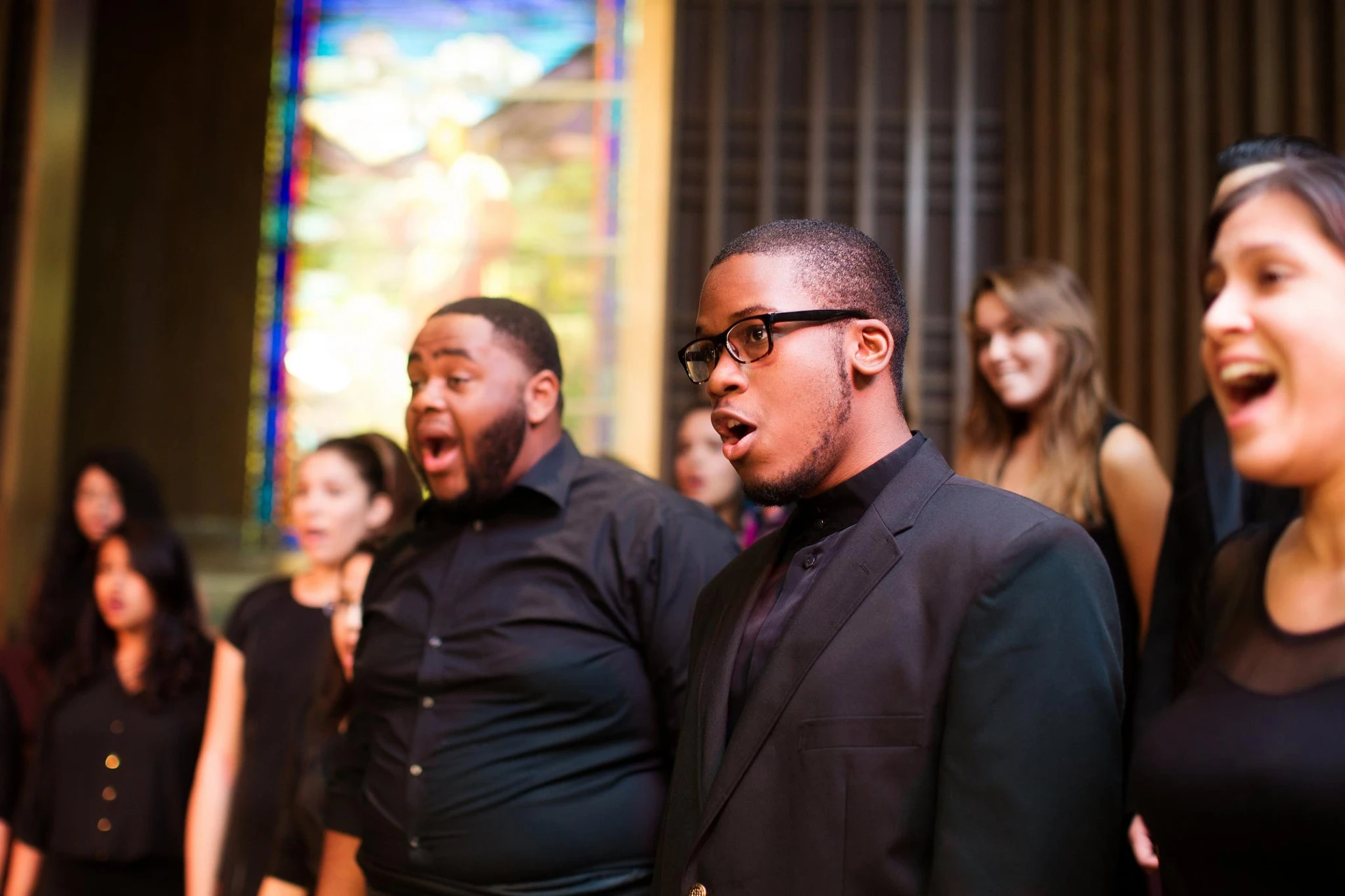 A young man, dressed in all black, join his peers as they look toward their director and sing