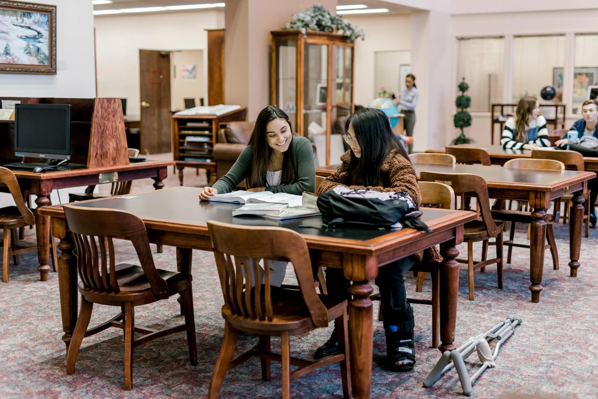 Two female, sit at a recentagular table at the library, looking through a shared textbook