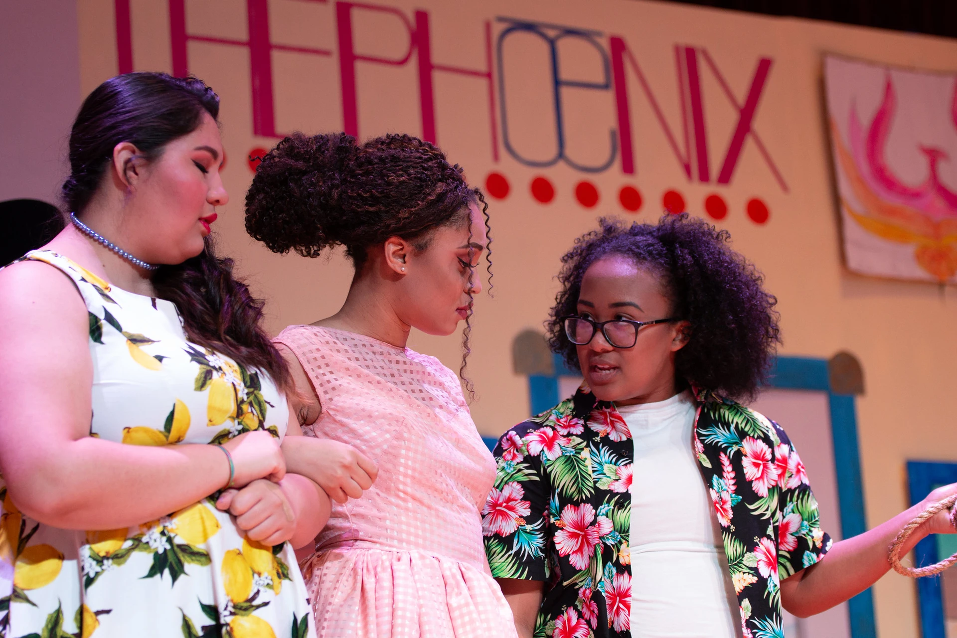 Three women, stand in front of their play background, and look at each other as the speak