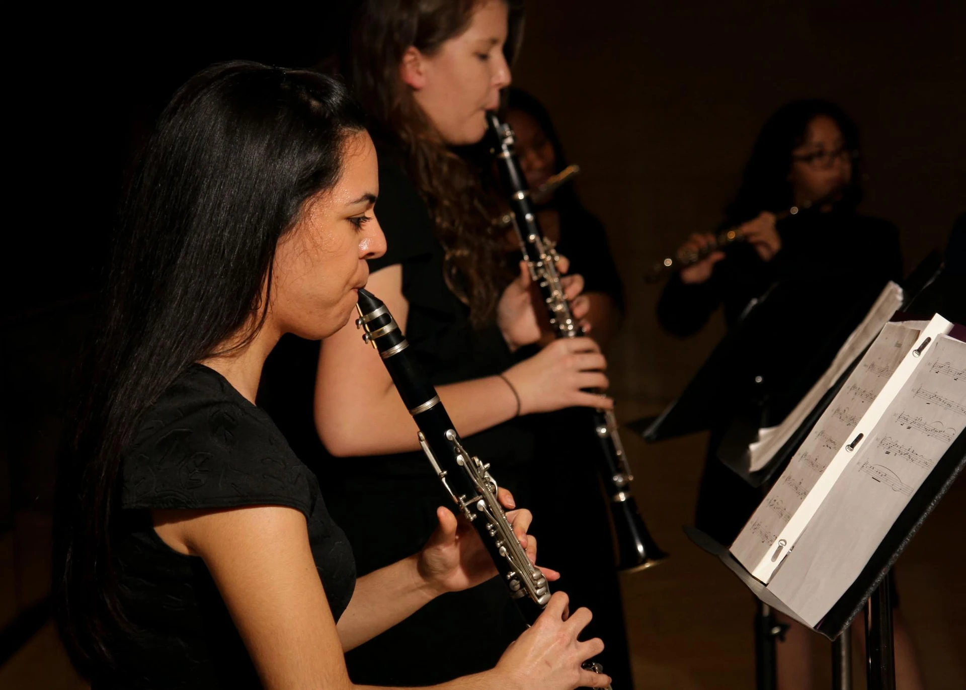 A clarinet player, with medium-lengthed dark hair, holds and plays her insturment while reading her music 