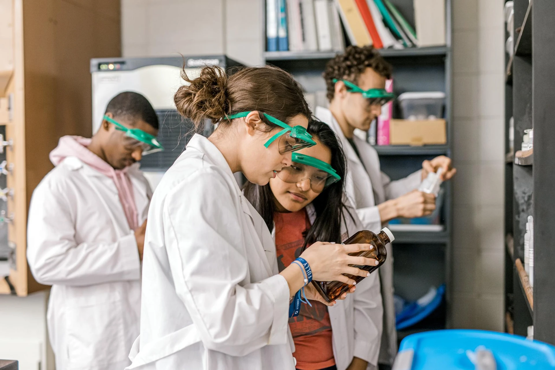 Two students, dressed in lab coats and goggles, pick up a bottle to read the label of the chemical