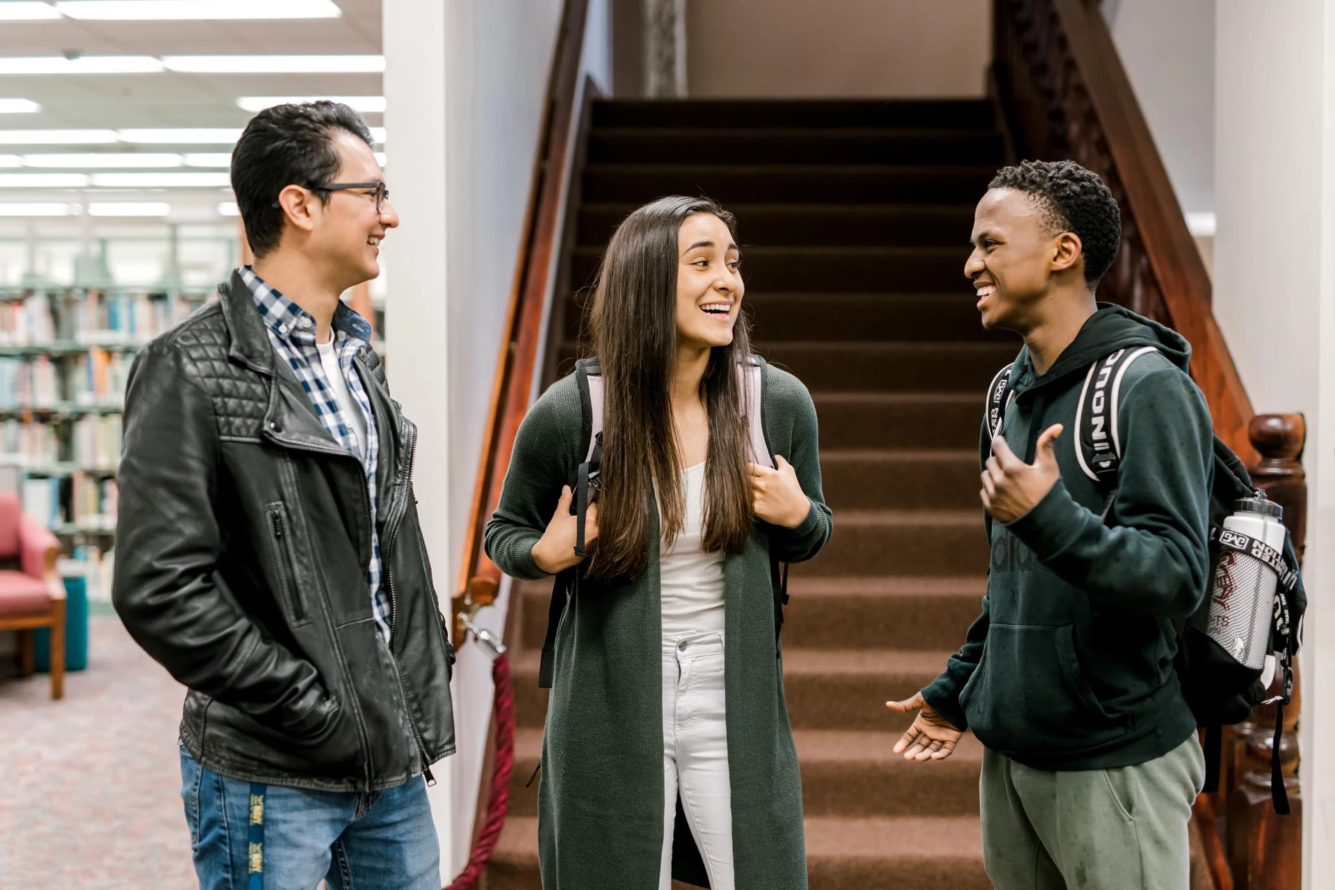 Three students stand in front of the carpeted-library stairs and laugh 