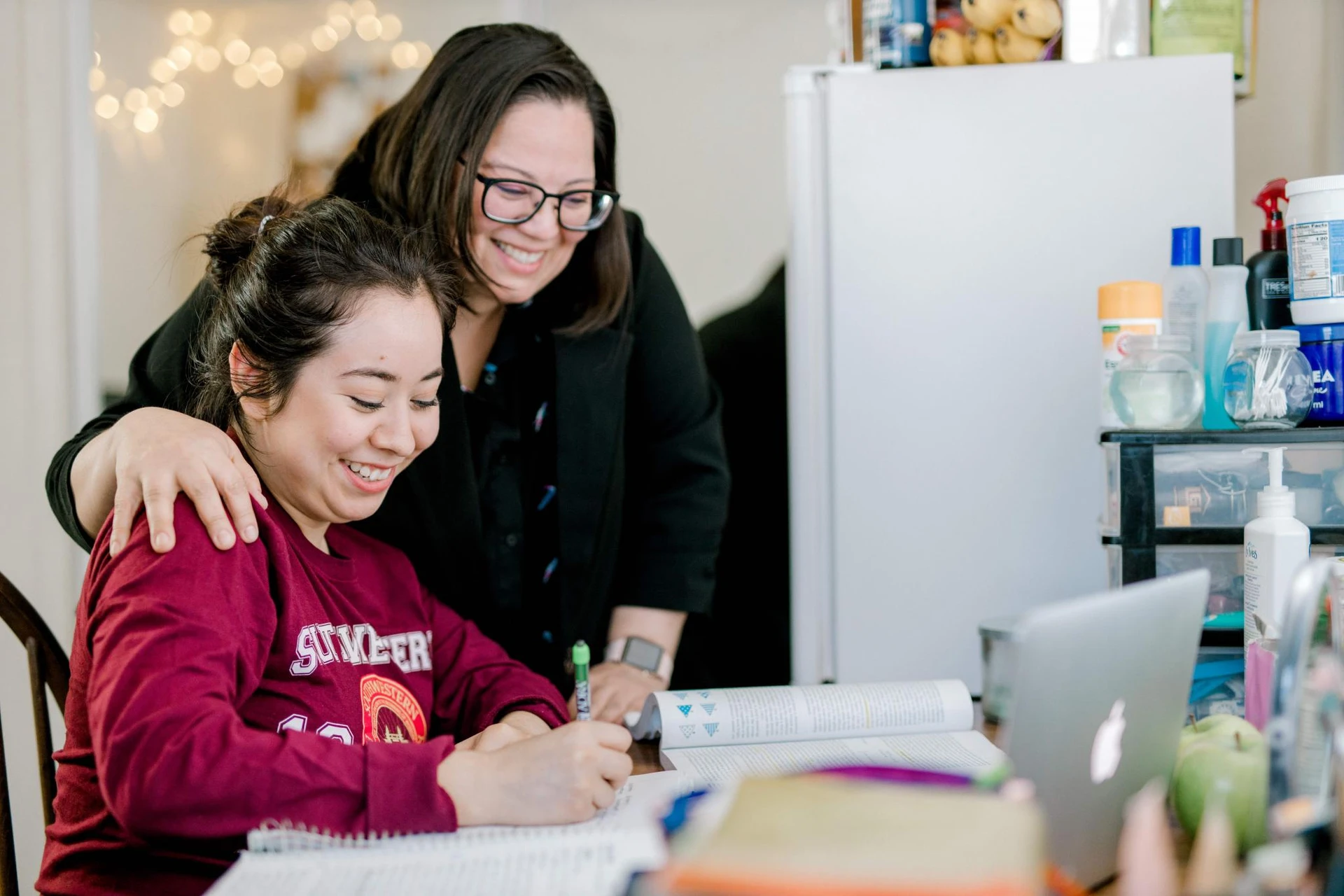 A student and her mother smile, both looking at a a computuer screen as the student does home work.