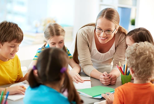 A teacher squats down and smiles as her students sit at a table and complete their homework